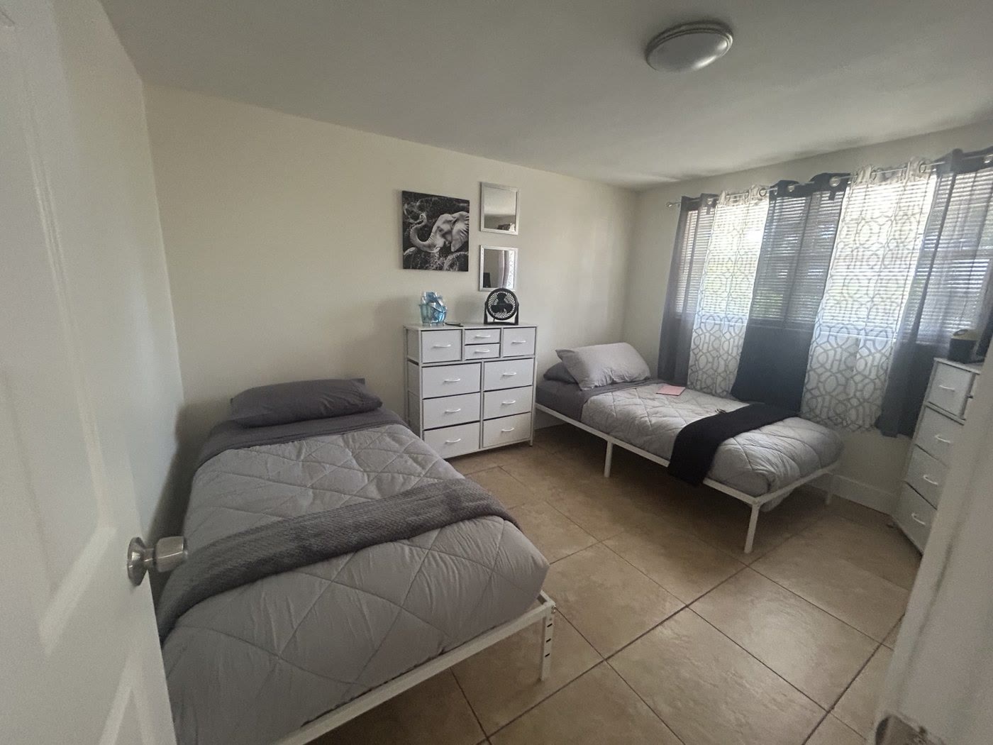 A bedroom with two made twin beds, gray bedding, a white dresser and a window with curtains.