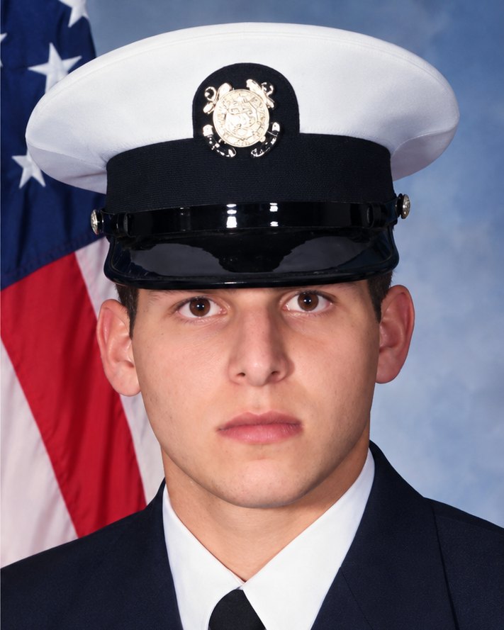 George's Coast Guard portrait in dress blues and a white cap, with the American flag behind him